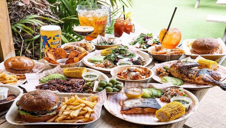 A wooden table is filled with various dishes including burgers with fries, grilled corn on the cob, fresh salads, bowls of olives, and roasted chicken. Drinks like iced tea and cocktails are also present, all set in a lush outdoor setting.