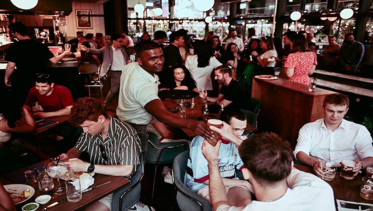 A bustling bar scene with diverse patrons enjoying drinks and socializing. A waiter, wearing a light green shirt, is serving a drink to a customer at a crowded table. The atmosphere is lively with people engaged in conversation and laughter.