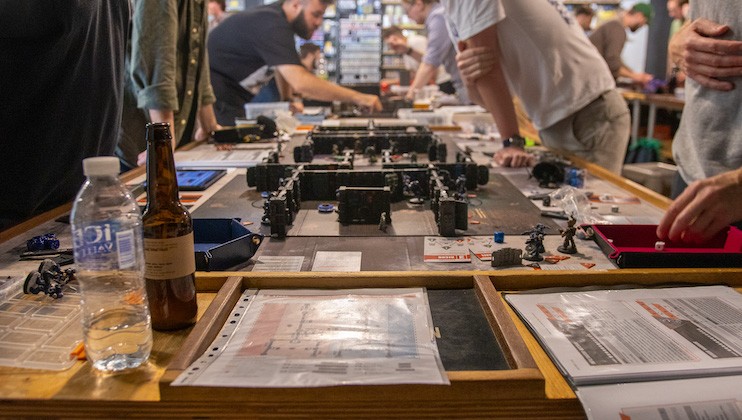 A crowded tabletop gaming event is depicted. People are gathered around large tables filled with game boards, miniatures, rulebooks, and gaming paraphernalia. A bottle of water and a beer bottle are visible in the foreground among the gaming materials.