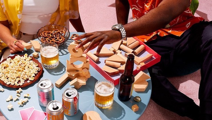 Two people play a block-stacking game on a table with various snacks and drinks. Items include popcorn, pretzels, beer, cans of soda, and wooden blocks. One person reaches for a block, while the other sits nearby. Bright and colorful setting.