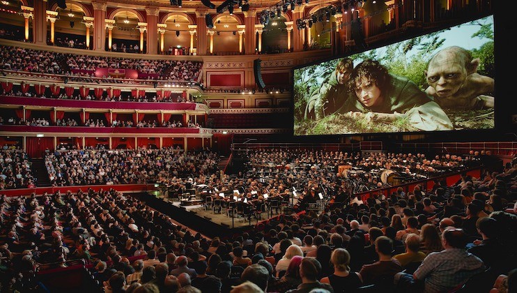 A large audience fills an opulent concert hall with multiple balconies. An orchestra performs onstage, and a giant screen above them displays a scene from a movie featuring two characters in a forest. The hall is illuminated with warm lighting.