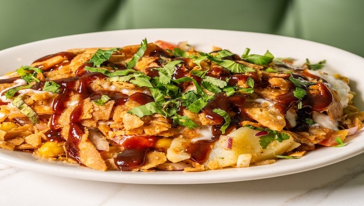 A white plate filled with chilaquiles, topped with tortilla chips, shredded cheese, sour cream, fresh cilantro, and drizzled with dark and creamy sauces. The background is slightly out of focus, with a hint of a green booth.