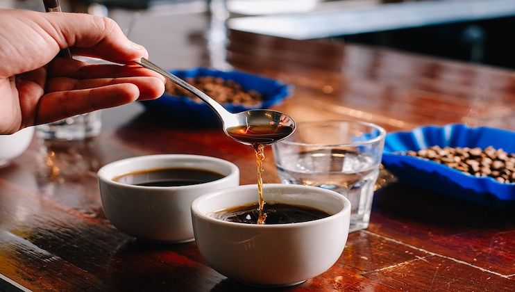 A hand holds a spoon dripping coffee into one of two white cups filled with coffee on a wooden table. In the background, there are blue trays with coffee beans and a glass of water. The setting appears to be a coffee tasting or cupping session.