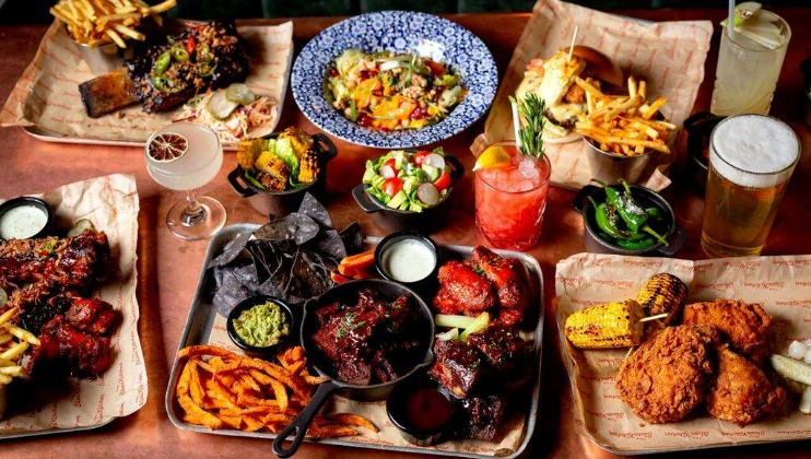 A table laden with various dishes including fried chicken, ribs, guacamole with blue corn chips, salads, fries, grilled corn, a variety of colorful cocktails, and a beer. The food is arranged on parchment-covered trays, creating a vibrant and appetizing spread.