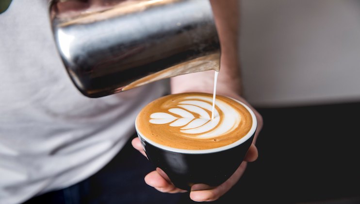 A person pours steamed milk in a pattern into a cup of coffee, creating latte art. The design features a heart-shaped pattern with rippling layers, demonstrating a barista's skill. The cup is black with a white interior, and the person's hand is visible holding it.