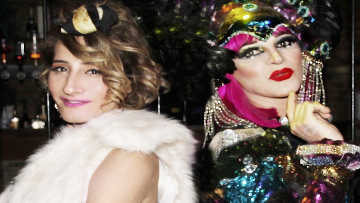 Two people posing together, one on the left has short, wavy hair, a fur shawl, and a pink top, while the other on the right is wearing vibrant, elaborate makeup, colorful sequined clothing, and a headdress. Both are looking at the camera with confident expressions.