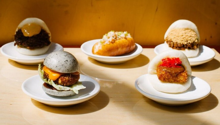 A variety of steamed bao buns are displayed on white plates. Each bun is filled with different ingredients, including crispy chicken, grilled meat, and various colorful vegetables and sauces. The background is a warm yellow, enhancing the vibrant presentation.