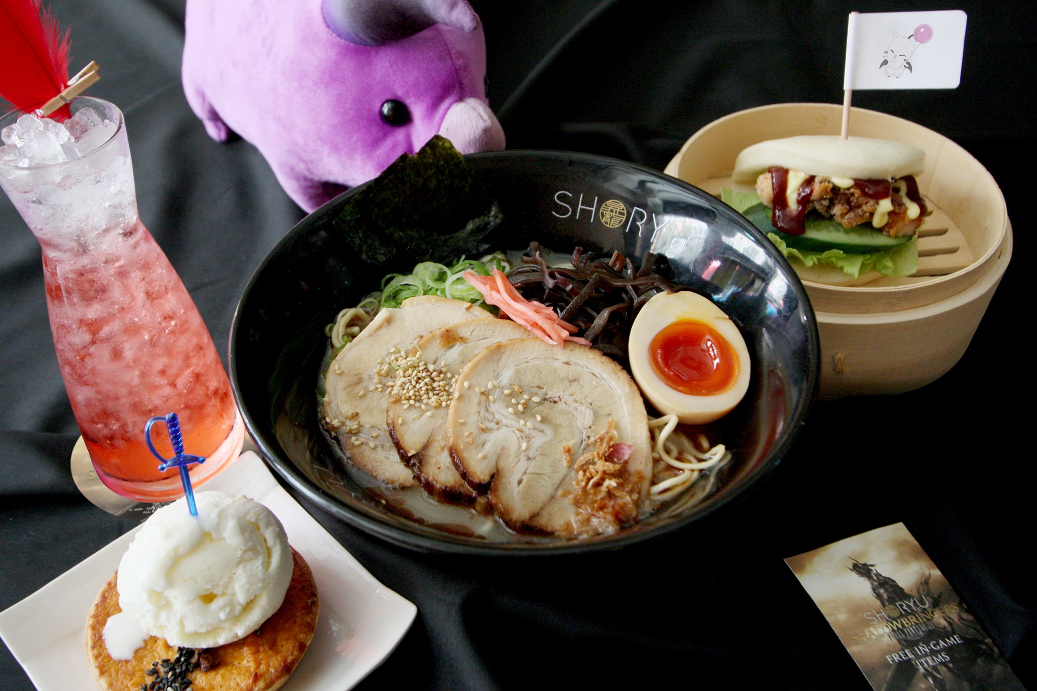 A bowl of ramen with sliced pork, half an egg, and vegetables is placed next to a steamed bun sandwich on a wooden plate. A pink drink with a red feather stirrer and a dessert topped with ice cream are also present. A plush purple toy bull peeks in from the background.