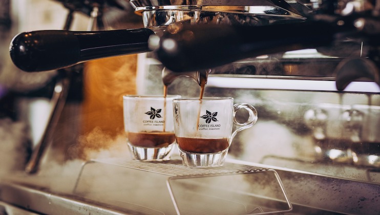 Two espresso shots being poured from a coffee machine into clear glass cups. Steam rises around the machine as the dark liquid fills the cups, creating a cozy, warm ambiance. The cups feature a logo with a simple flower design and the text Coffee Island.