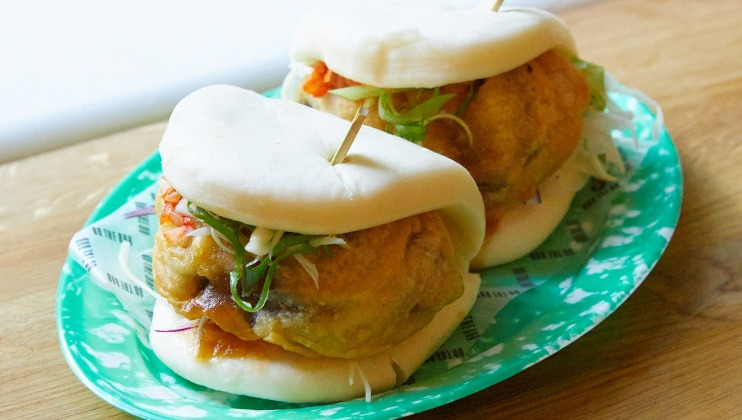 Two steamed bao buns filled with crispy battered fried chicken, shredded vegetables, and sauce held together with toothpicks, served on a green and white patterned plate.