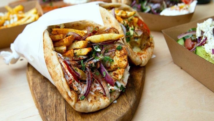 A close-up of a delicious gyro sandwich wrapped in pita bread, filled with sliced grilled meat, seasoned french fries, red onion, tomatoes, and fresh herbs. The sandwich is placed on a wooden serving board, with more food items in cardboard containers in the background.