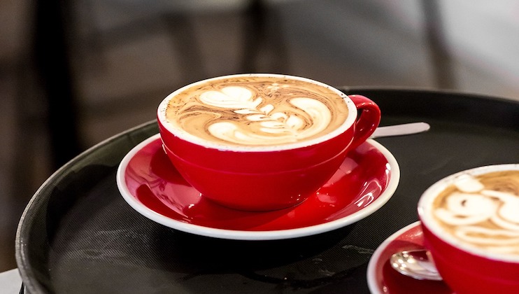 A red cup filled with a cappuccino or latte featuring intricate latte art, placed on a matching red saucer. The cup is on a black tray. Another similar cup is partially visible in the foreground. The background is blurred out.