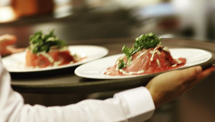 A server in a white shirt carries a tray with two plates of a gourmet dish. Each plate features a neatly arranged pile of thinly sliced red meat, topped with green leafy garnish and drizzled with sauce. The background is blurred, highlighting the food.