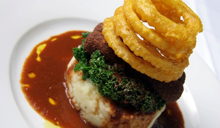 A gourmet dish features mashed potatoes topped with a layer of green kale, browned meat, and a stack of golden onion rings. The dish is artfully presented, drizzled with rich brown gravy on a white plate.