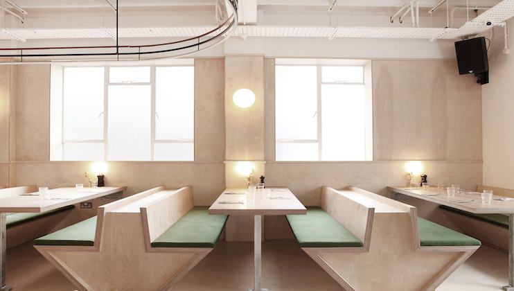 A minimalist cafe interior featuring light wood paneling and green cushioned booth seating. Two large windows let in natural light, while modern pendant lights add ambiance. Tables are set with menus and utensils, creating a clean and inviting atmosphere.