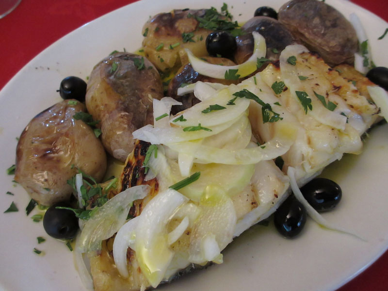 A plated dish featuring a grilled white fish fillet topped with onions and chopped herbs, surrounded by boiled potatoes and black olives. The dish is presented on a white plate with a red background.