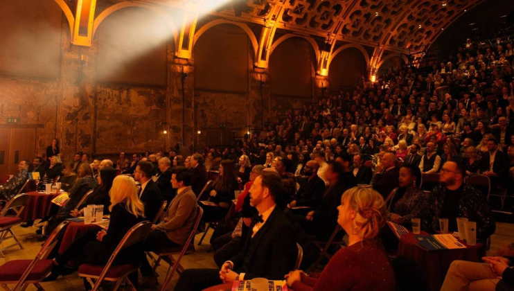 A diverse group of people seated in a grand, ornately decorated theater with arched ceilings and intricate wall designs. A spotlight illuminates part of the audience, who are attentively watching something off-frame. The atmosphere is formal and anticipatory.