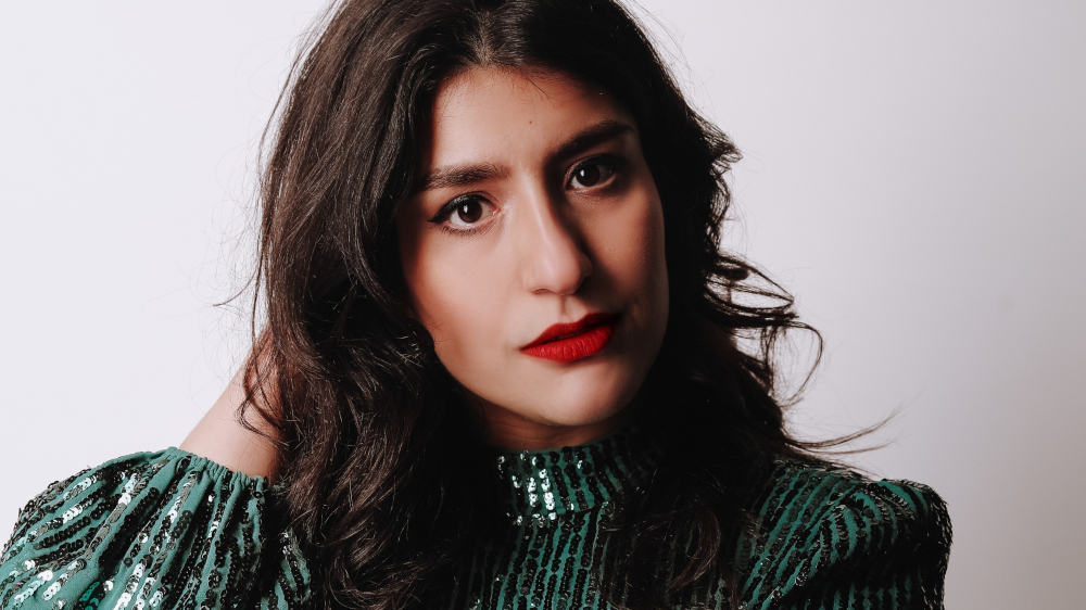 A woman with long dark hair wearing a green sequined top poses against a plain white background. She has a serious expression, bold red lipstick, and her left hand is touching her hair.