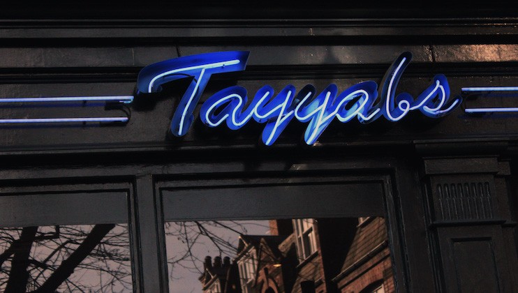 A neon blue sign displaying the name Tayyabs is mounted on a dark storefront. Reflections of buildings and tree branches are visible in the storefront windows below the sign.