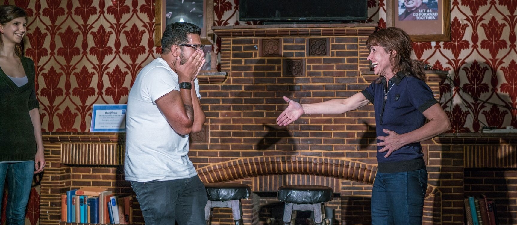 Three people are standing in front of a brick fireplace. A man in a white shirt is laughing with his hands over his mouth, facing a smiling woman in a navy shirt who has her hand extended for a handshake. A third person in a green top is standing in the background, smiling.