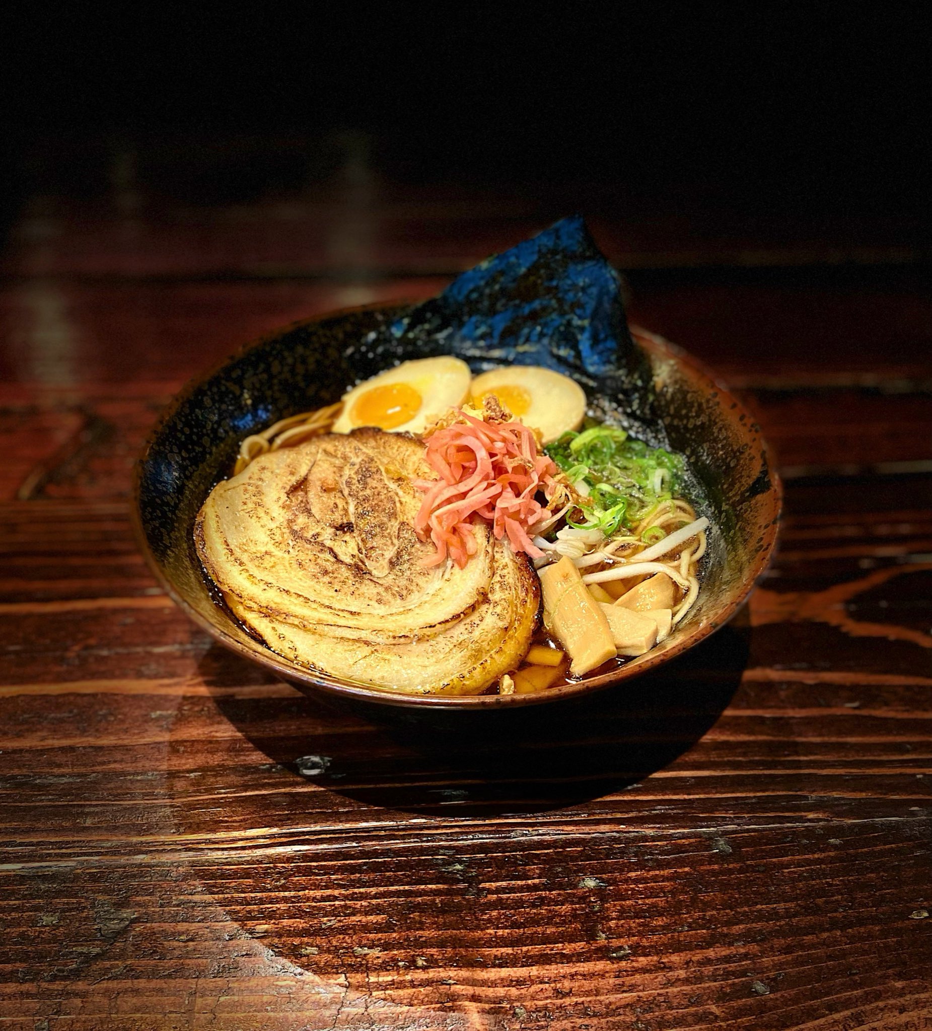 A bowl of ramen is displayed on a dark wooden surface. The ramen features slices of pork, a halved soft-boiled egg, green onions, bamboo shoots, pink pickled ginger, and a sheet of nori. The broth and noodles are visible under the toppings.