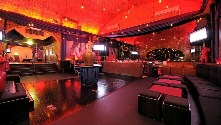 An interior of a dimly lit nightclub featuring a high arched ceiling, red and orange lighting, multiple flat screen TVs, and modern seating areas with black cushions and red tables. Art sculptures and decorative elements add to the stylish ambiance.