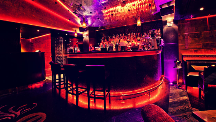 A dimly lit bar with red and purple lighting. The curved bar counter features several high stools and is lined with various bottles and glasses. The ceiling has intricate designs, and pendant lights add to the cozy ambiance. Tables and chairs are seen in the background.