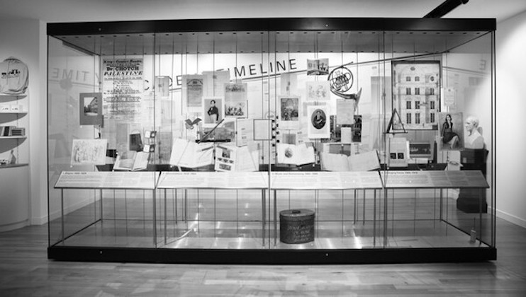 A black and white image displays a glass museum exhibit case containing an array of historical artifacts, documents, framed photos, and various items. The display is well-lit and meticulously arranged, inviting viewers to explore the historical timeline presented within.