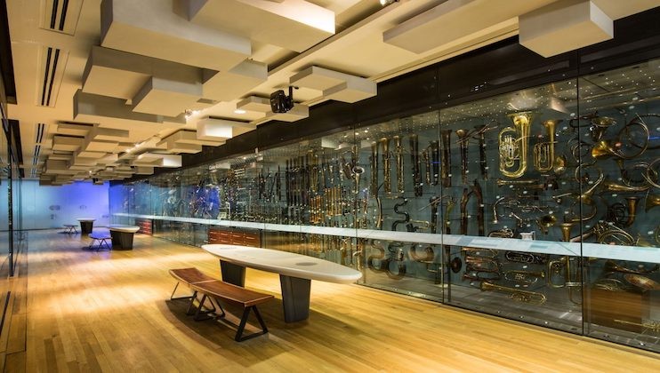 Museum exhibit with a glass wall displaying a variety of brass and wind instruments. The room features wooden flooring, modern seating benches, and ceiling acoustic panels. The instruments are meticulously arranged and well-lit for viewing.