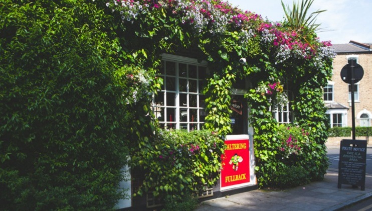 A quaint building covered with lush green vines and vibrant flowers stands on a sunny day. A red sign near the door displays Charrington Fullback. A chalkboard menu is visible on the sidewalk, enhancing the inviting atmosphere of the establishment.