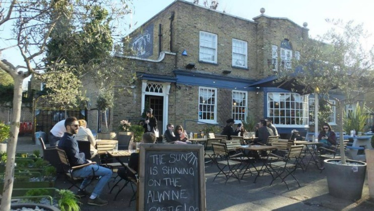 People are sitting and chatting at outdoor tables in front of a rustic brick pub with large windows. The pub has a blue door and trim, and plants in pots decorate the patio. A chalkboard sign reads THE SUN IS SHINING ON THE ALWYNE CASTLE.