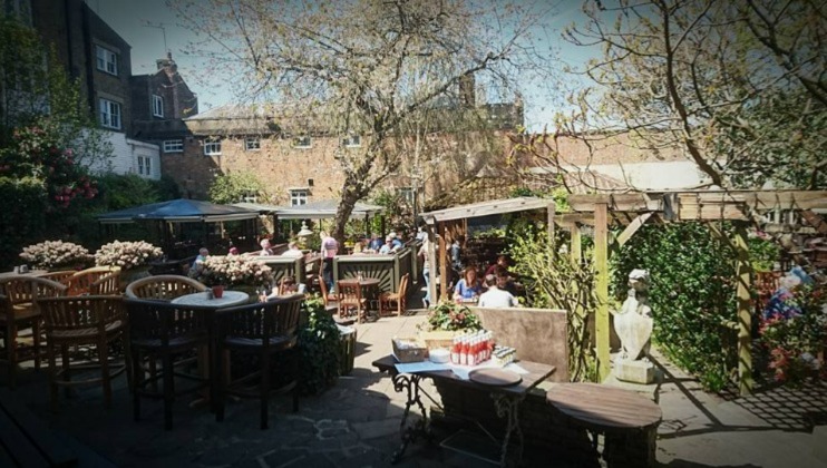 A sunlit outdoor garden patio with several wooden tables and chairs. People are seated, engaged in conversation. The area is surrounded by lush greenery, including trees and potted plants, creating a cozy and inviting atmosphere. Some buildings are visible in the background.