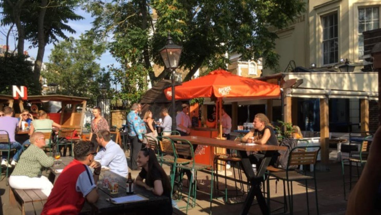 A lively outdoor bar scene with people sitting under orange umbrellas and at tables, enjoying food and drinks. The area is shaded by trees, and the atmosphere is relaxed and social on a sunny day.