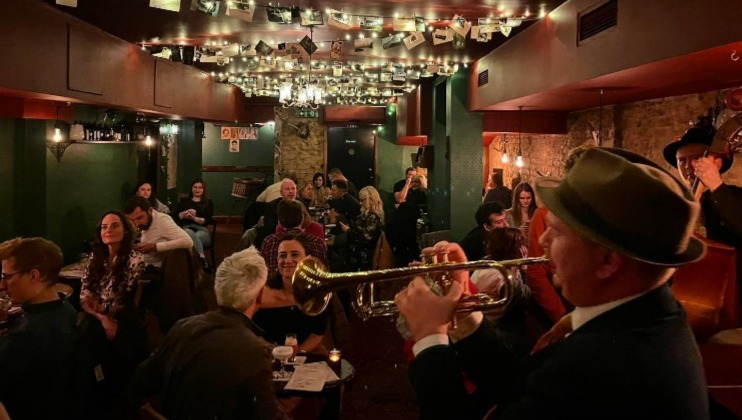 A lively, warm-lit jazz bar with patrons seated at tables, enjoying the music. In the foreground, a man in a hat plays the trumpet passionately. The ceiling is adorned with small lights, and the room has a cozy, intimate atmosphere, with people chatting and enjoying the performance.