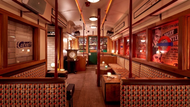 A cozy, dimly-lit restaurant interior designed to look like a vintage train carriage. Booths with patterned upholstery line both sides, a clock is attached to one wall, and retro posters adorn the walls. Soft lighting creates an inviting and nostalgic atmosphere.