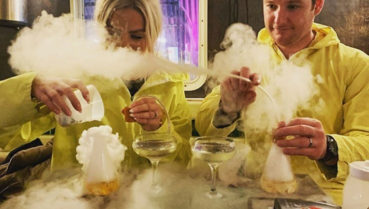 Two people in yellow jackets are creating smokey drinks in a bar. They are using various tools to add ingredients to the glasses, which are emitting clouds of white smoke, creating a dramatic, misty effect. The scene appears lively and experimental.