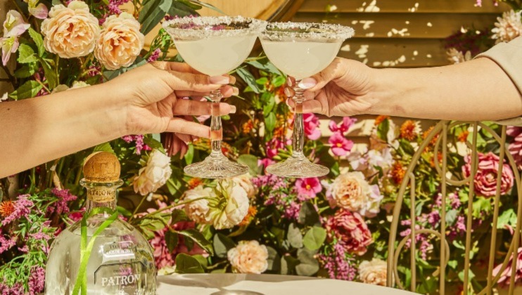 Two hands clink margarita glasses with salted rims in a celebratory toast. The background features a vibrant and colorful garden adorned with various flowers. A bottle of Patron tequila is placed on the table.