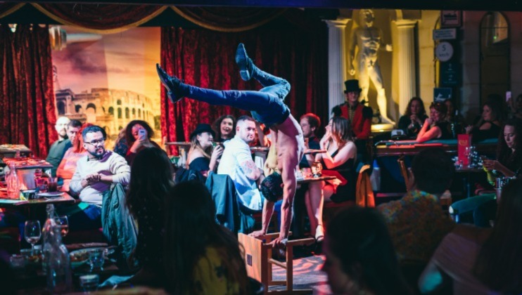 A shirtless performer balances on one hand in the middle of a dimly lit, crowded cabaret with red velvet curtains and a statue in the background. The audience, seated at tables, watches the acrobatic act attentively.