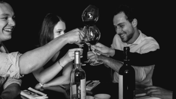 A group of people sit at a table clinking their wine glasses in a toast, surrounded by bottles and tableware. The image is black and white, capturing a moment of camaraderie and celebration.