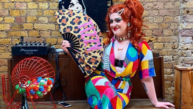 A person with red hair styled in waves, adorned with colorful hairpins, and wearing bright makeup, sits on a table. They hold a leopard print fan and wear a vibrant, multi-colored outfit. Next to them is a bingo cage and a DJ mixer with a brick wall backdrop.