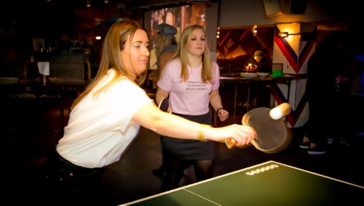 Two women engaged in a game of table tennis in a lively indoor setting. The woman in the foreground is about to hit the ball with her paddle while the other watches attentively. The background is filled with dim lighting and various people and decorations.