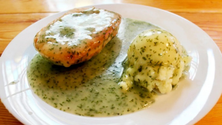 A plate of food featuring a baked potato with a creamy herb sauce and a side of mashed potatoes also topped with the same creamy herb sauce, all set on a white plate atop a wooden table.