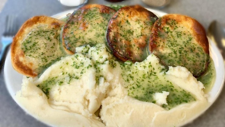 A plate filled with mashed potatoes and four pies drenched in green parsley sauce. The creamy mashed potatoes are topped with a drizzle of the same green sauce, creating a cohesive, appetizing appearance.