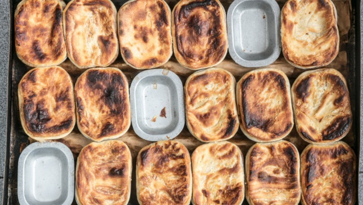 A baking tray filled with several small, rectangular pies, most of which have golden brown crusts. Some pies have slightly darker, charred spots on their surfaces, while a few pie dishes appear empty. The pies are arranged closely together in neat rows.