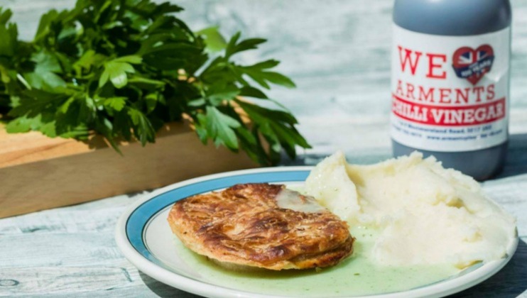 A plate with a pie and mashed potatoes is positioned in the foreground, with a bottle of chili vinegar to the right and a bunch of parsley on a wooden board in the background. The setting appears to be on a light-colored surface.