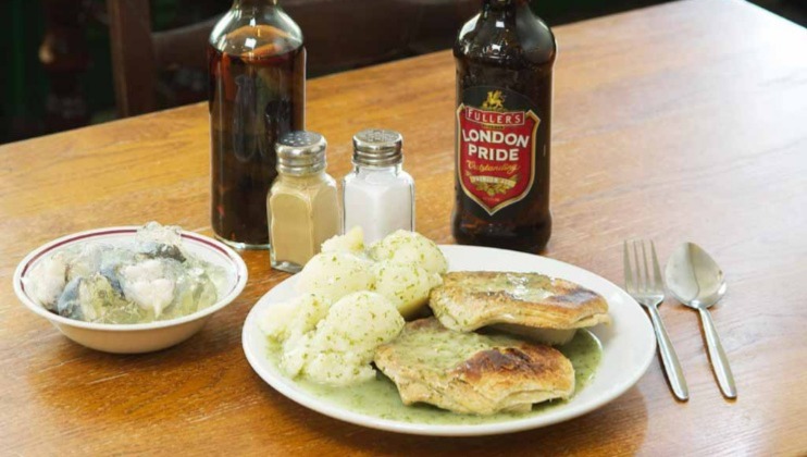 A meal on a wooden table with a plate of pie, mashed potatoes, and green sauce. Next to it are a bowl with a side dish, salt and pepper shakers, a brown sauce bottle, and a bottle of Fuller's London Pride beer. Fork and spoon are beside the plate.