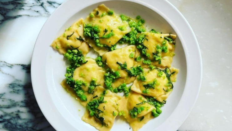 A white plate contains several square ravioli pieces covered with a green peas and basil sauce. The dish is presented on a marble surface.