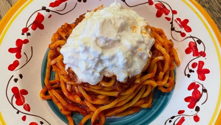 A plate of fettuccine pasta topped with a rich red tomato sauce and a large dollop of burrata cheese. The plate has a colorful floral pattern with red flowers and green stems around the rim. The dish is set on a wooden table.