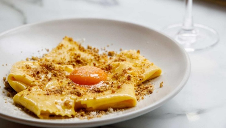 A plate of folded pasta squares garnished with a soft-cooked egg yolk in the center, and sprinkled with a brown crumbly topping, is served on a white plate. The plate rests on a marble surface with a wine glass visible in the background.