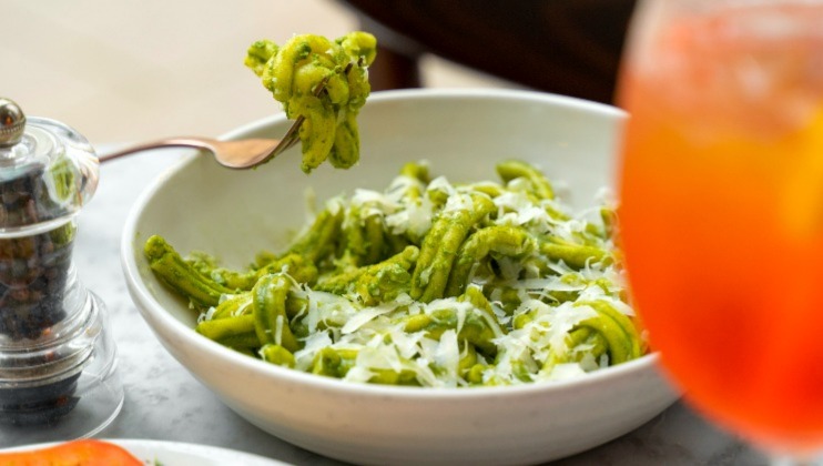 A bowl of pasta covered in green pesto sauce and sprinkled with grated cheese is positioned on a table. A fork holds up a bite of the pasta. Nearby, a clear pepper grinder and a glass containing a bright orange drink can also be seen.